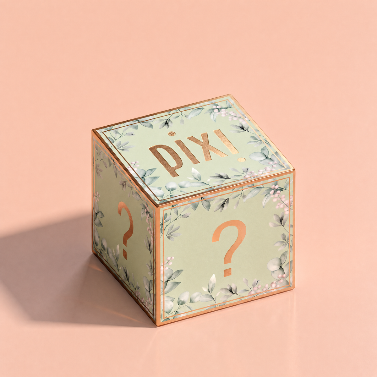 A decorative cube-shaped box with a pale green floral print, metallic rose-gold trim, and 'pixi' on the top face, photographed on a soft peach-pink background.