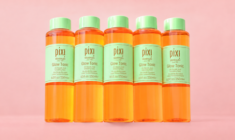 Five Pixi Glow Tonic bottles filled with bright orange liquid, arranged in a row against a solid pink background. View 2