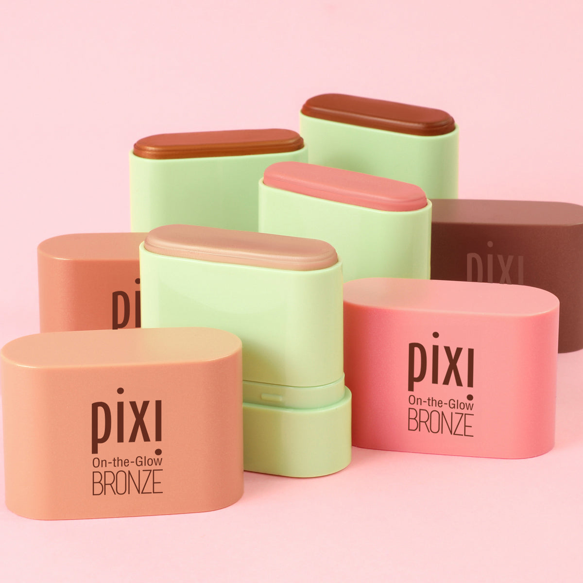 Six Pixi On-the-Glow Bronze stick blush compact cases in various shades of peach, pink, and light green, arranged on a solid pink background.