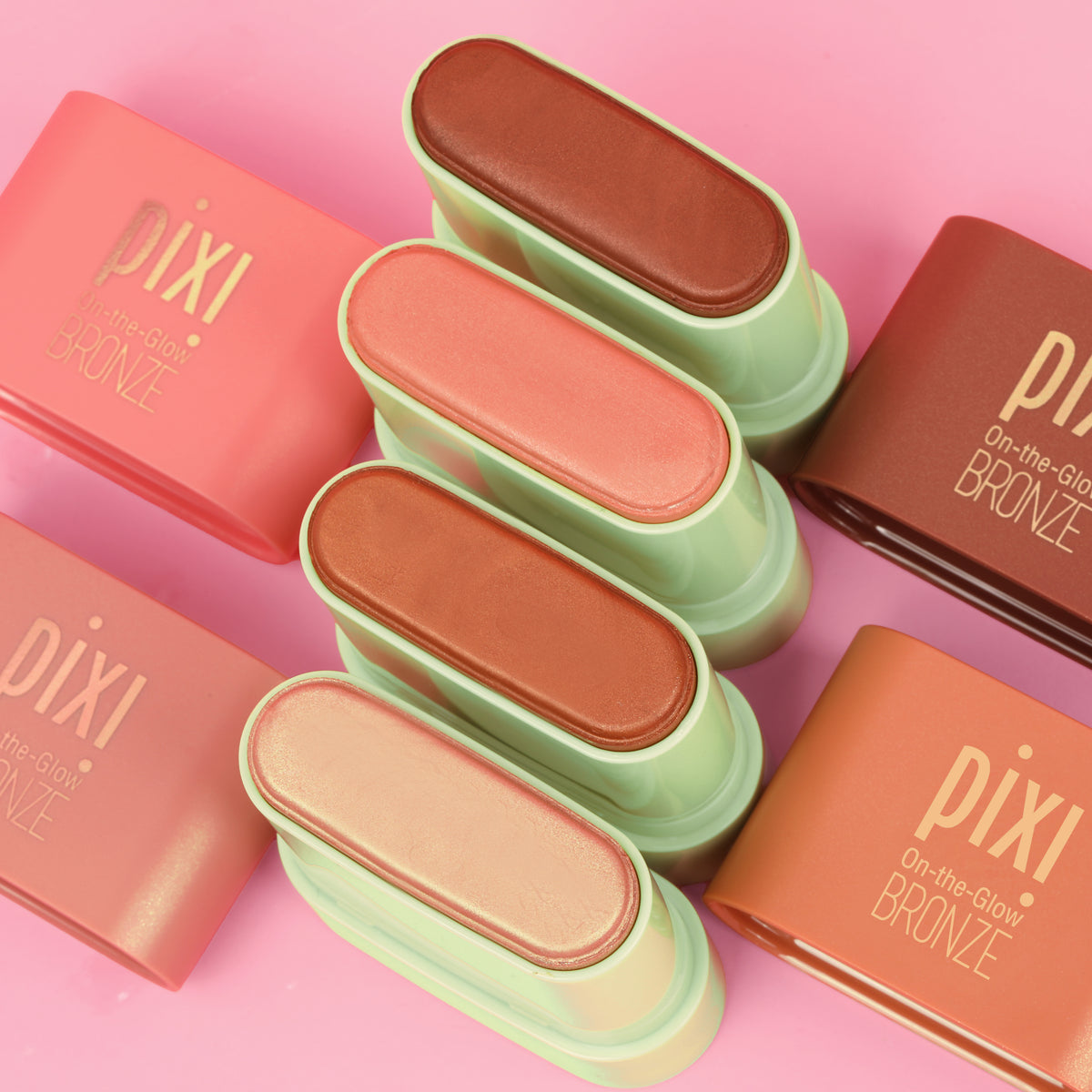 Six Pixi On-the-Glow Bronze face powders in various warm shades, arranged vertically in the center with their coral-pink and terracotta-colored rectangular boxes on a solid pink background.