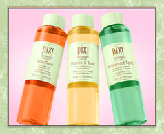 Three Pixi SkinTreats toner bottles arranged in a fan shape against a pink background with a decorative green border. The bottles contain orange, yellow, and green liquids and are labeled Glow Tonic, Vitamin-C Tonic, and Antioxidant Tonic.