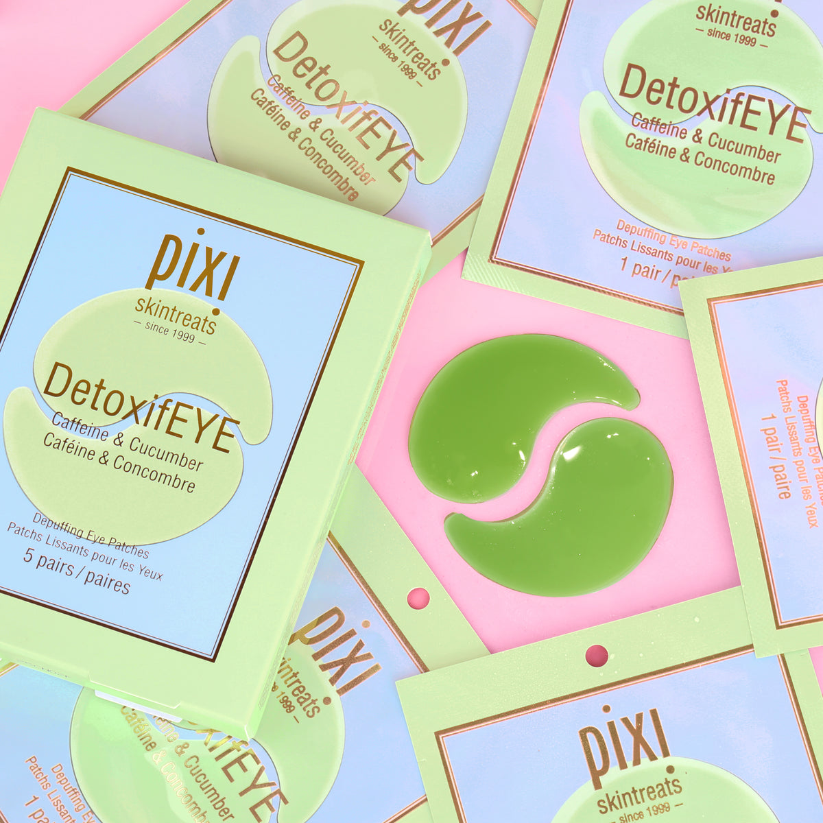Several Pixi Skintreats DetoxiEYE caffeine and cucumber eye patches and their packaging are arranged on a pastel pink background. The packaging is light green and lavender, and one green patch is shown.
