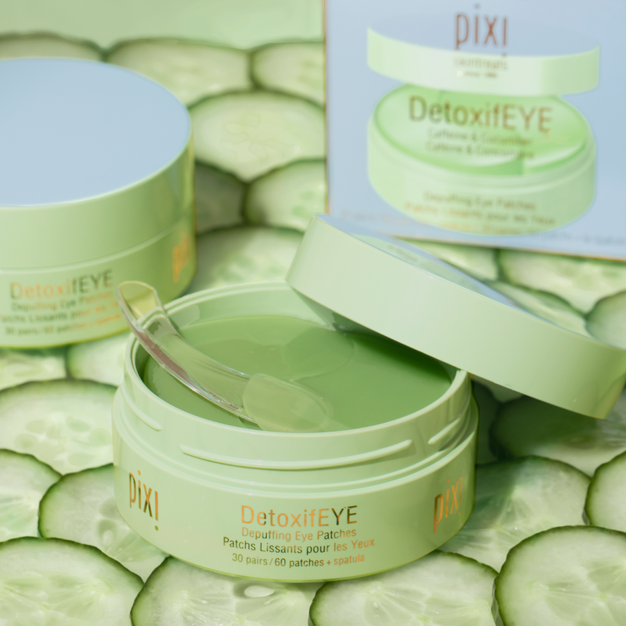 Pixi DetoxifEYE lifting eye patches shown in an open jar with a clear applicator spoon, alongside a closed jar, its packaging box, and sliced cucumber props on a light surface.