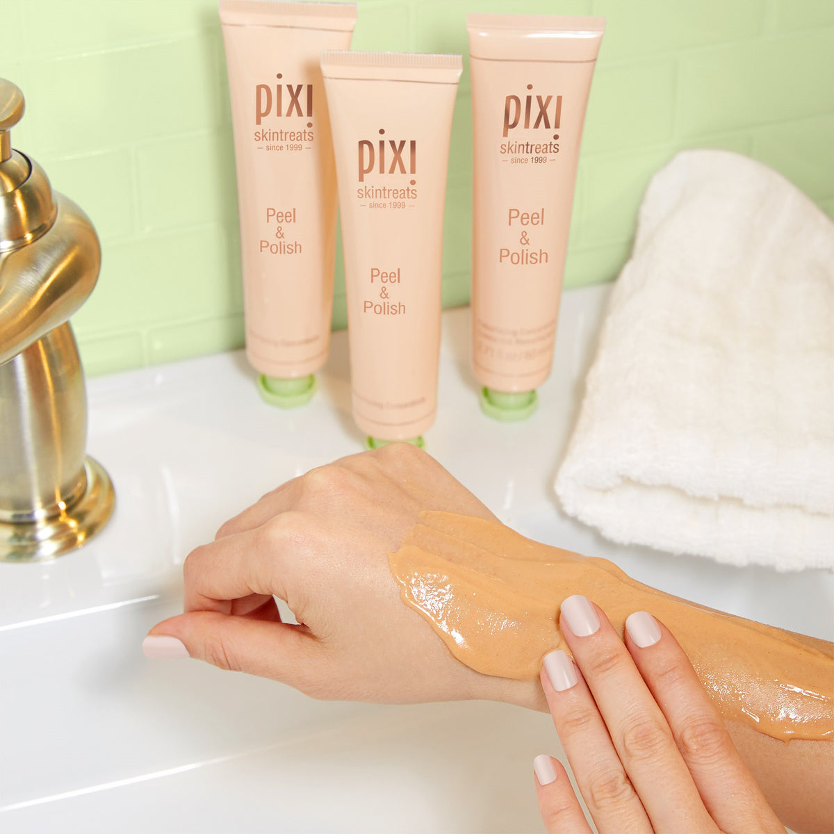 Three tubes of pixi skin treats Peel & Polish cream are lined up on a bathroom counter next to a folded white towel, with a hand showing the thick, glossy cream product applied to the forearm.